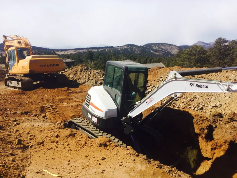 Mini excavator digging a deep hole in red soil, a larger excavator and foothills behind it