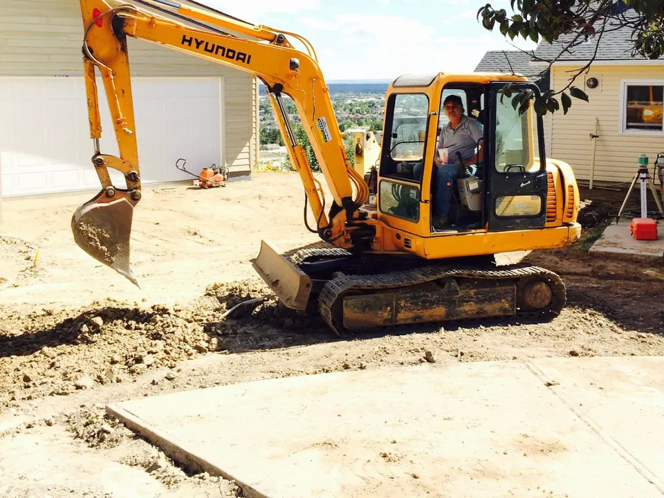 Mini excavator digging along a new concrete slab, laser level set up behind it