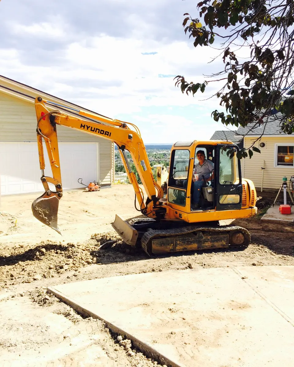 Operator in a Hyundai mini excavator digging along a new concrete slab, laser level set up behind it