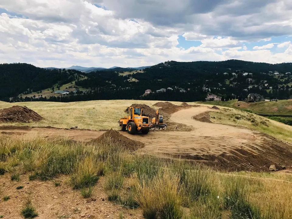 Wheel loader and excavator cutting a building pad and drive on an open foothills lot under a cloudy sky