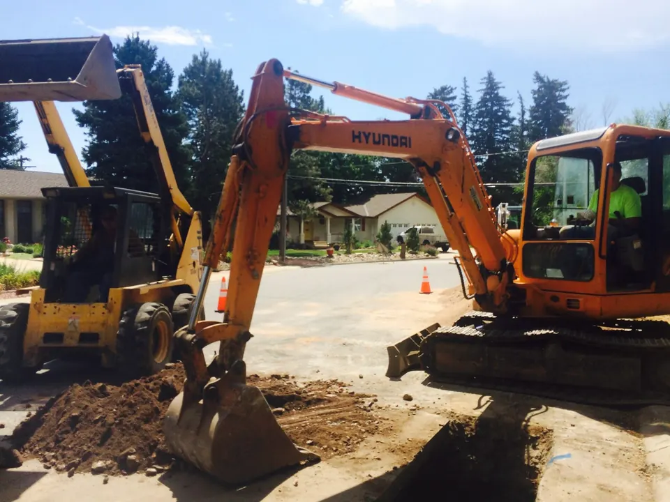 Orange mini excavator digging a trench across a residential street, cones and a skid steer beside it