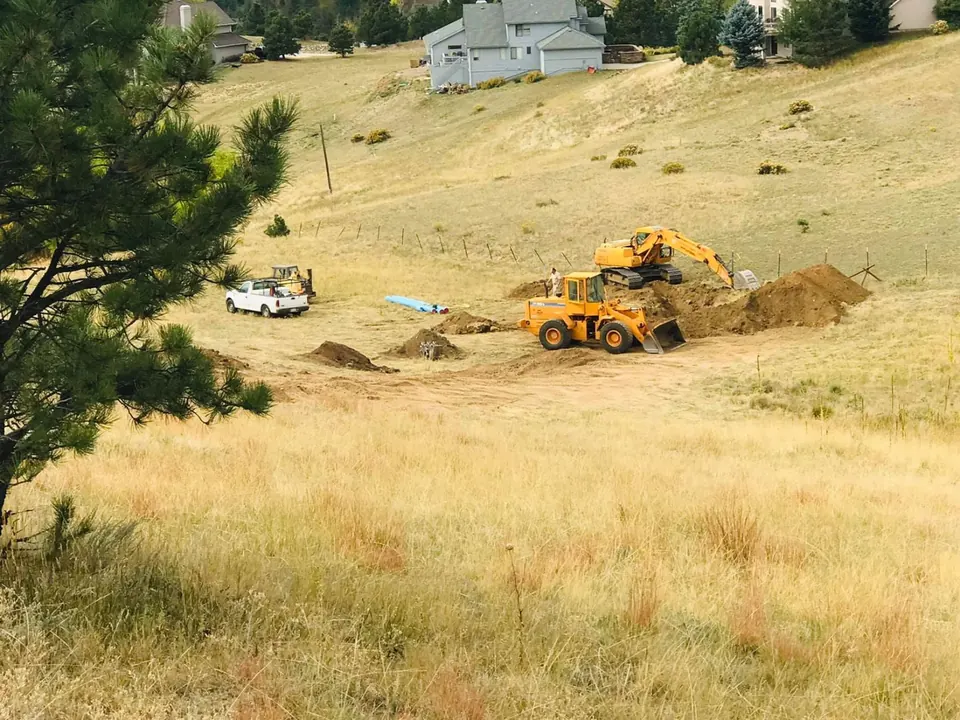 Wheel loader and excavator trenching on a grassy hillside lot, blue water pipe laid out nearby