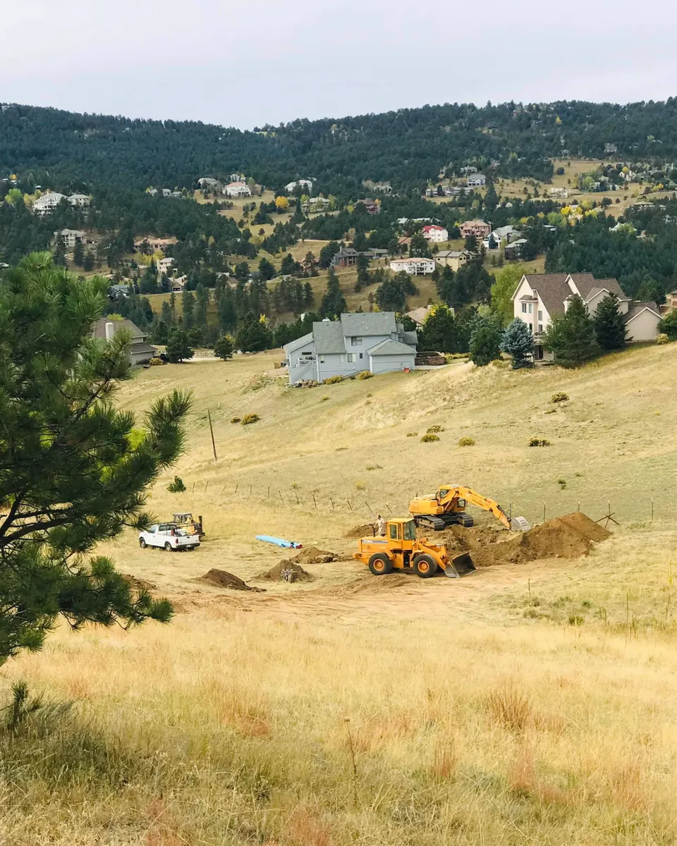 Loader and excavator trenching on a grassy hillside lot, blue water pipe laid out nearby
